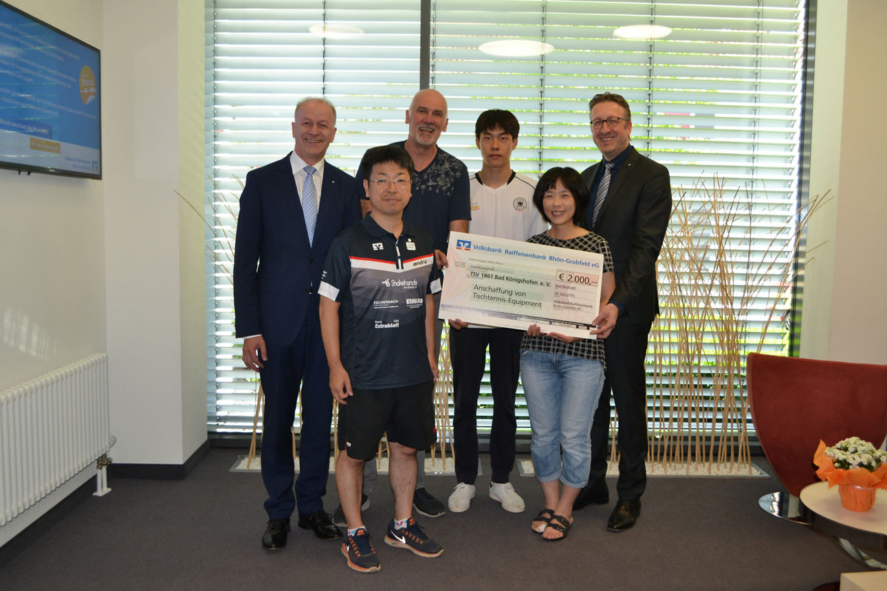 Fünf Männer und eine Frau stehen vor einem Fenster. Die Frau hält einen Spendenscheck in der Hand