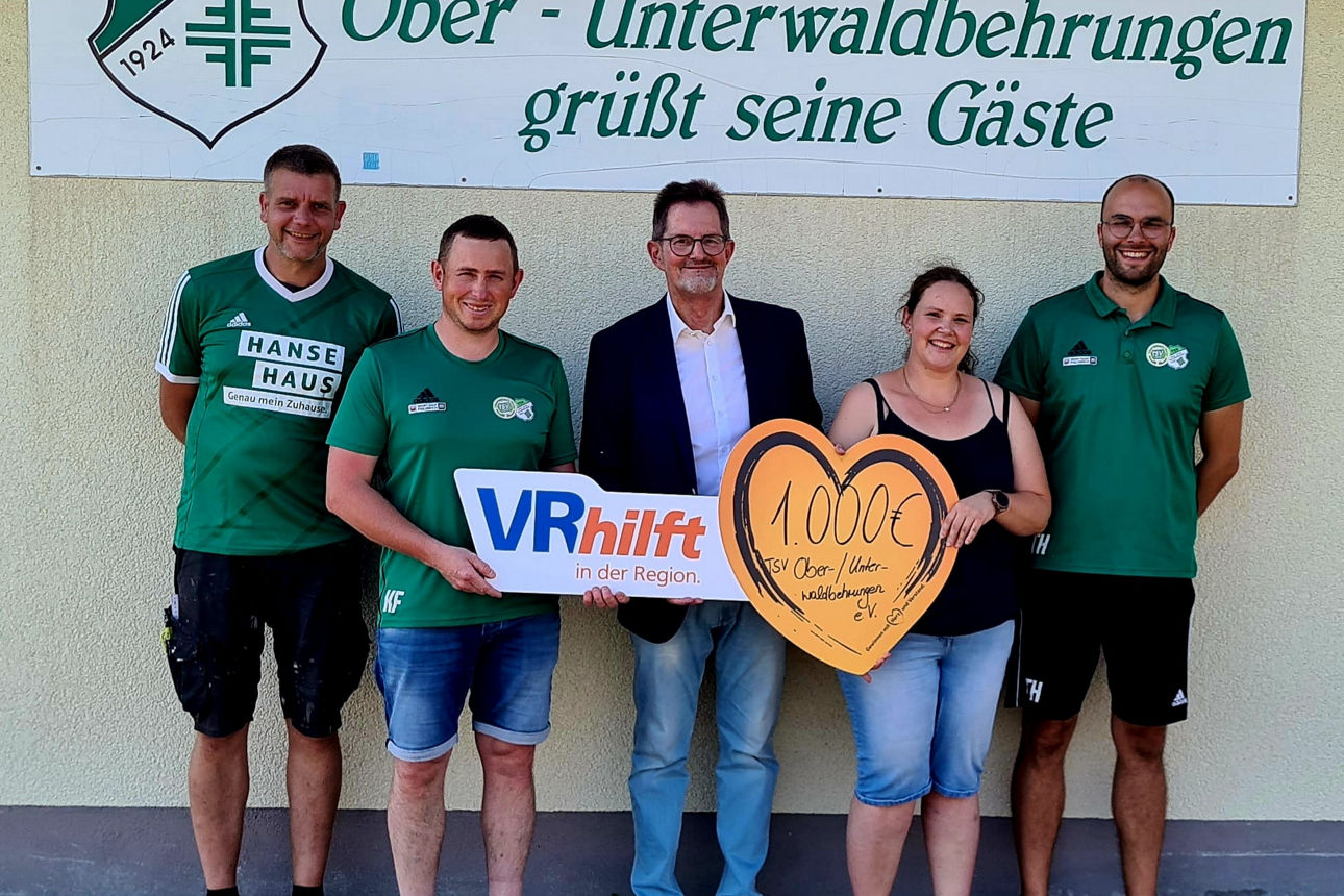 Fünf Personen stehen vor einer Wand mit dem Logo und Namen des TSV Ober-Unterwaldbehrungen, zwei von ihnen halten Schilder