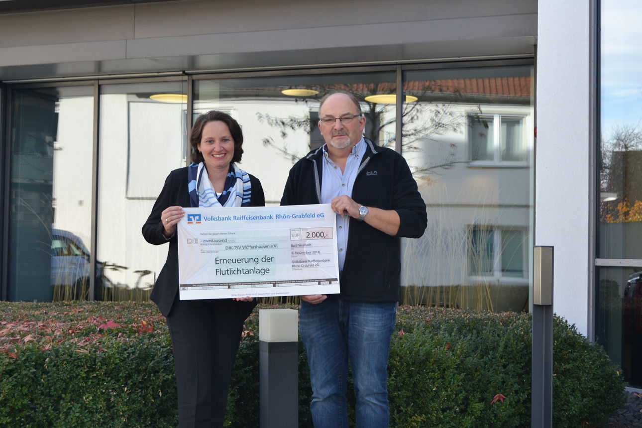 Ein Mann und eine Frau stehen vor einem Gebäude und halten einen Spendenscheck in der Hand.