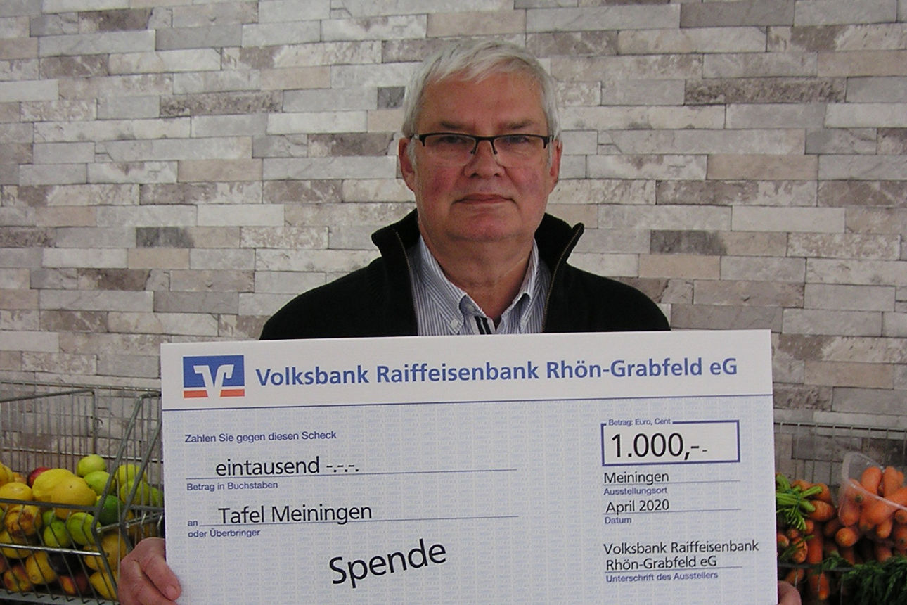 Ein Mann hält ein Spendenschild über 1.000 Euro für die „Tafel Meiningen“ vor Regalen mit Obst und Gemüse in den Händen 
