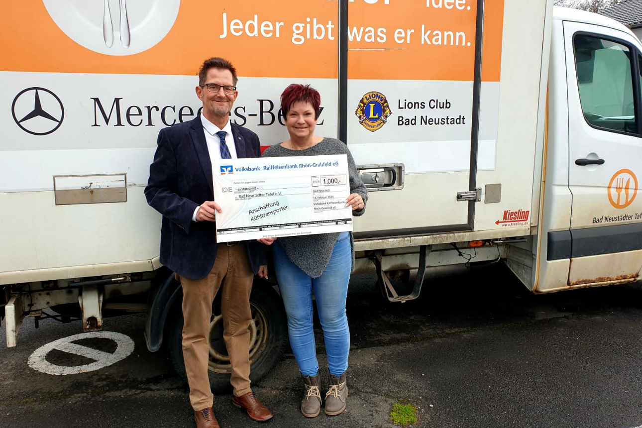 Ein Mann und eine Frau die vor einem Transporter der Tafel stehen halten einen Spendenscheck in der Hand
