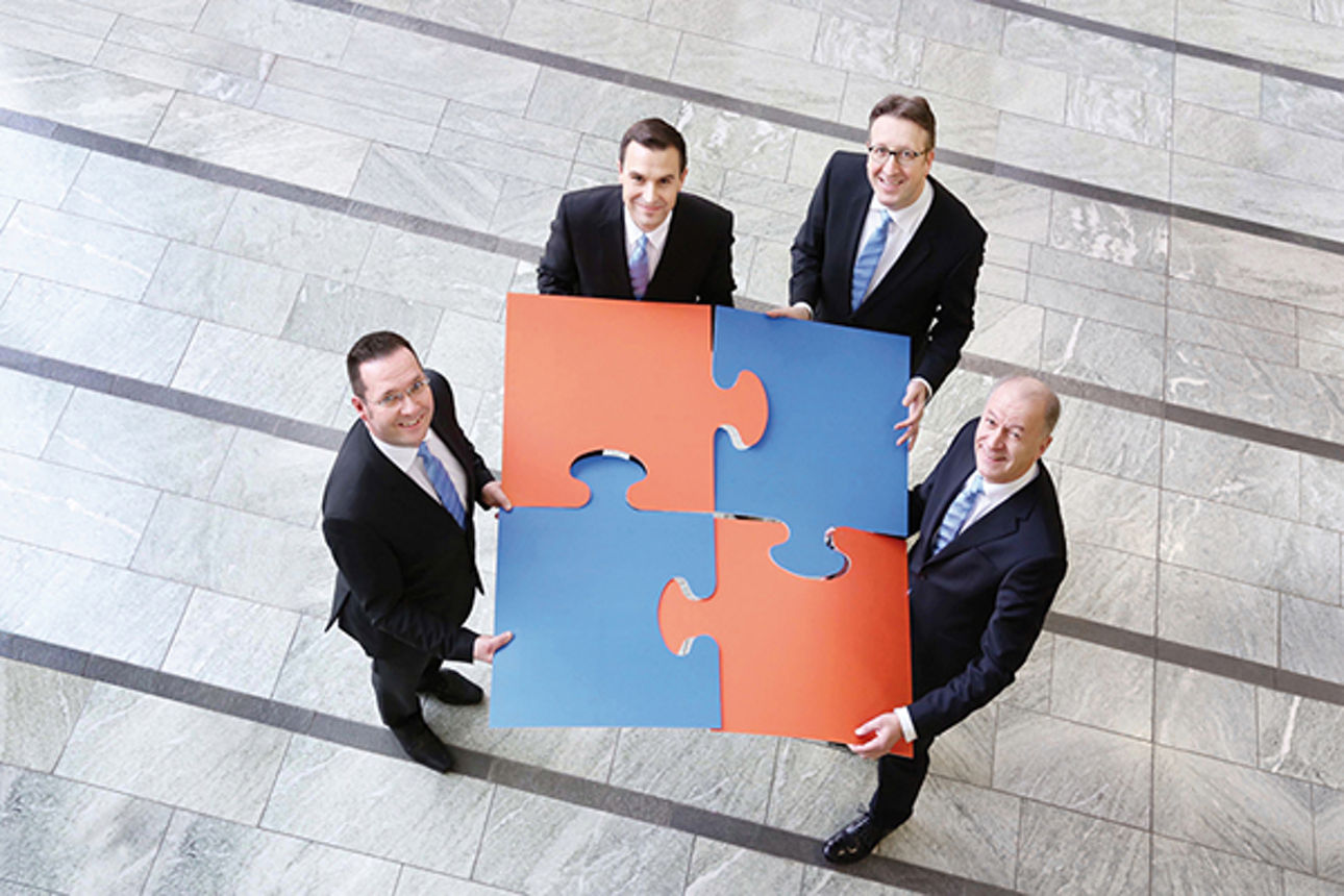 Vier Männer in Anzug schauen nach oben in eine Kamera und lächeln dabei, während jeder ein großes Puzzleteil hält 