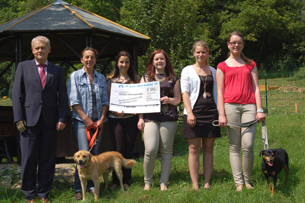 Sechs Personen stehen im Freien vor einem Pavillon eine Person in der Mitte hält einen großen Spendenscheck in der Hand.