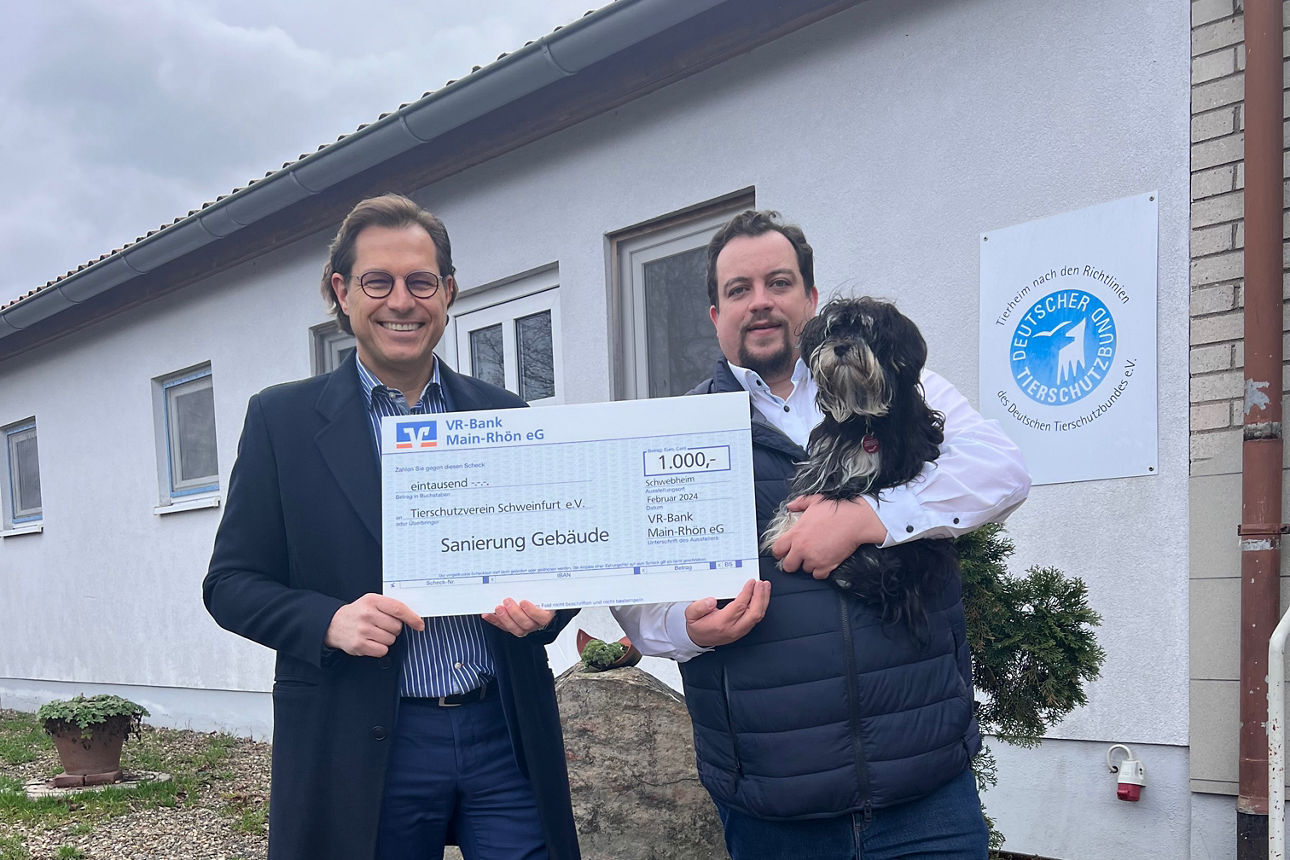 Zwei Männer stehen vor einem Gebäude, einer hält einen großen Scheck mit der Aufschrift „1.000 € Sanierung Gebäude“