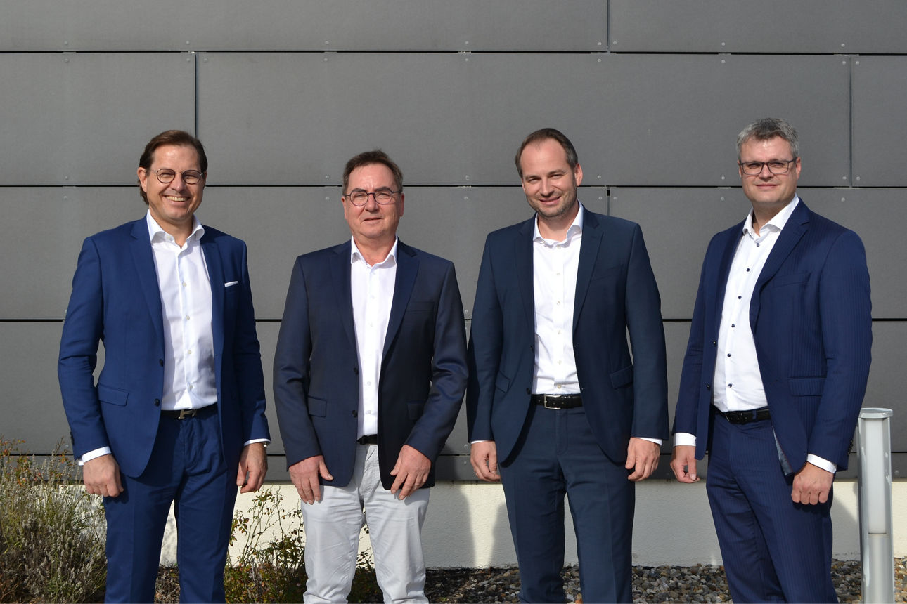 Vor einer grauen Hauswand stehen vier Männer, die Anzüge tragen 