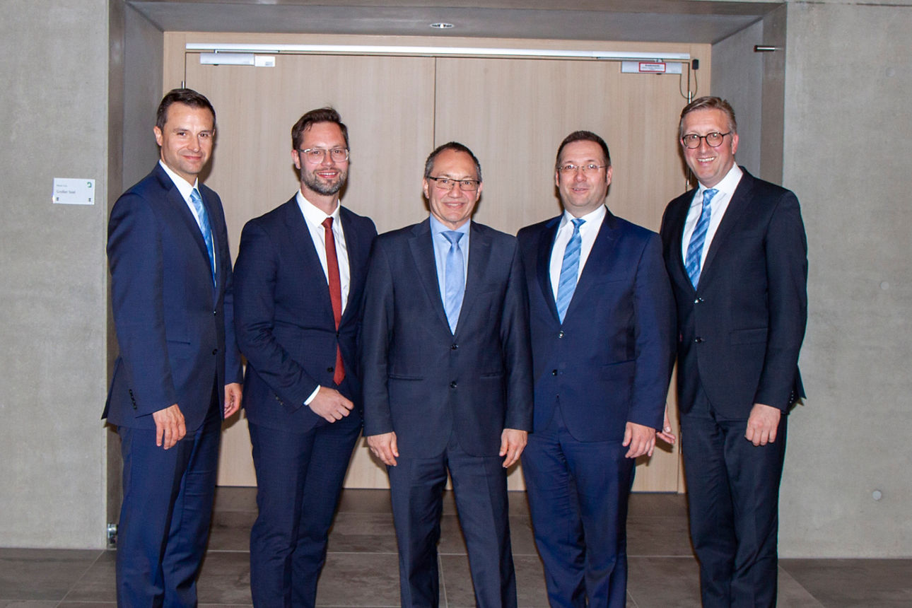 Vor einer Holztür stehen fünf Männer, die Anzüge tragen und in die Kamera lächeln 