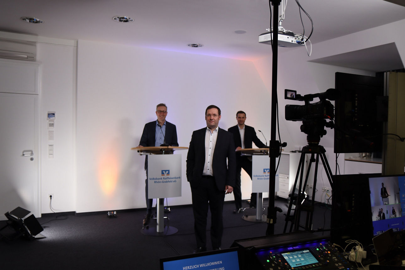 In einem Raum in dem viele Technikgeräte stehen sind drei Männer in Anzug zu sehen 