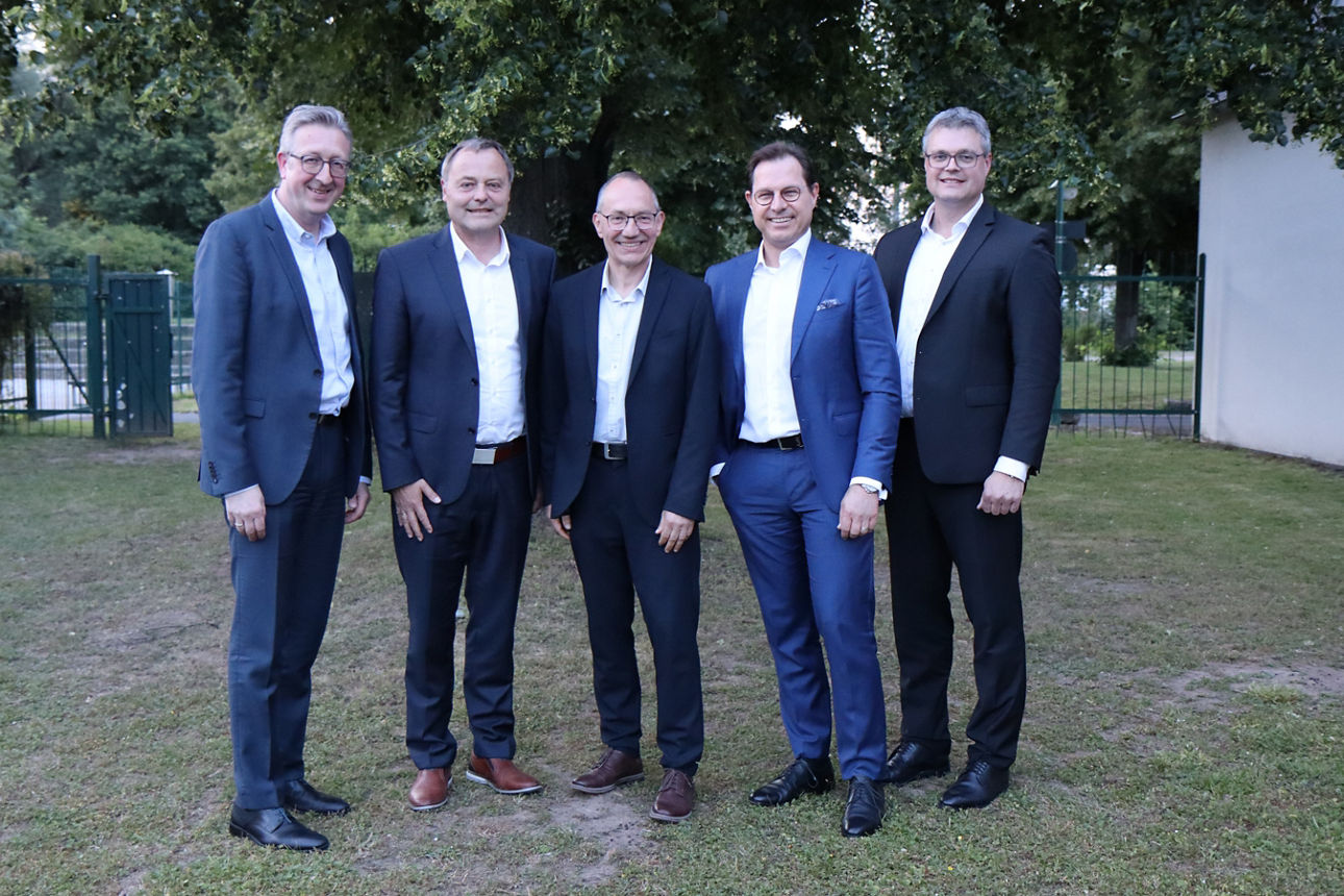 Im Freien stehen fünf Männer die Anzüge tragen und in die Kamera lächeln