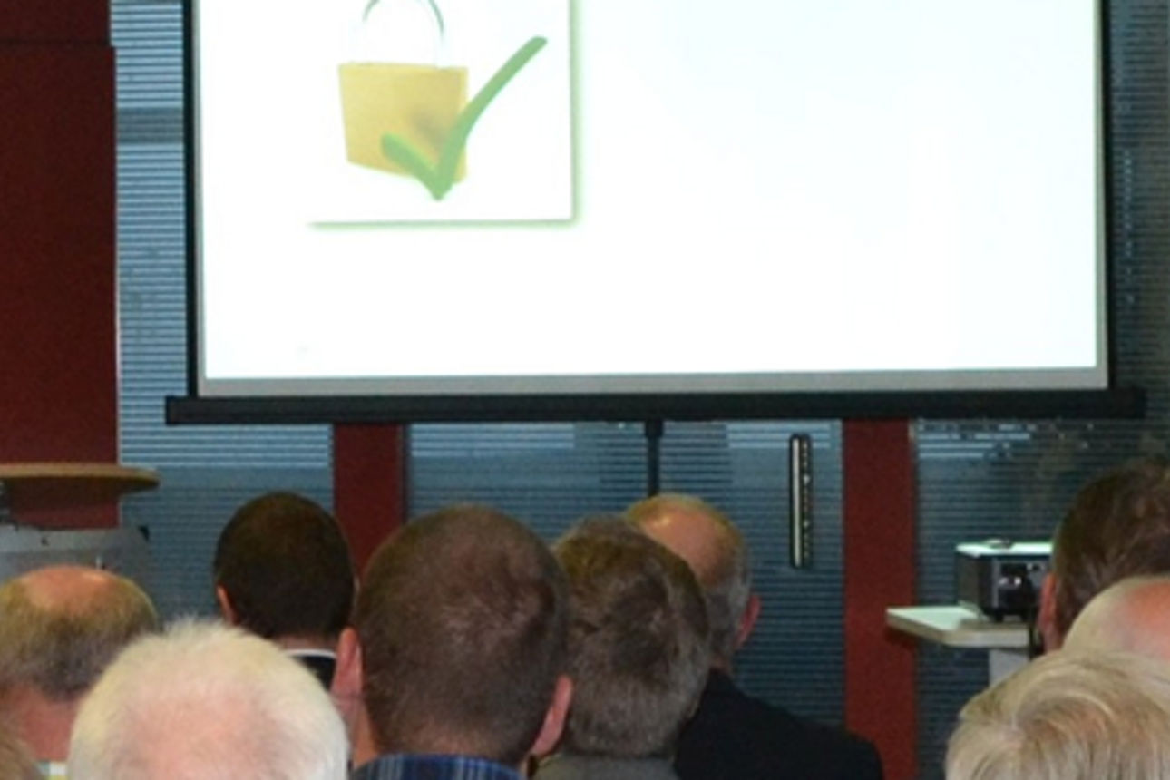 Ein Mann hält eine Rede an einem Rednerpult vor einem Publikum, auf einer Leinwand ist ein Symbol mit einem grünen Haken 