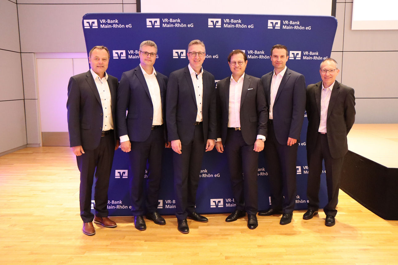 Vor einer blauen Wand in einer Halle stehen sechs Männer die Anzüge tragen und in die Kamera lächeln