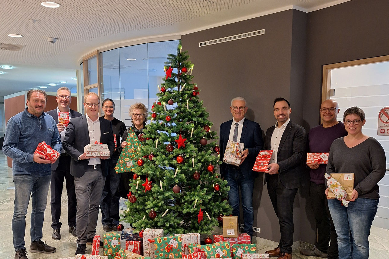 Einige Personen stehen mit Geschenken in der Hand um einen geschmückten Weihnachtsbaum. Vor diesem liegen weitere Geschenke