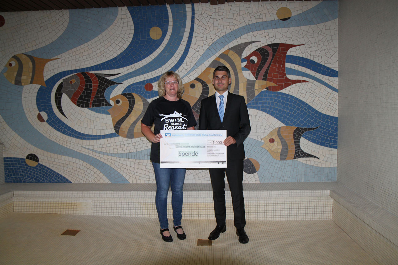 Ein Mann und eine Frau stehen vor einer Wand und halten einen Spendenscheck in der Hand 