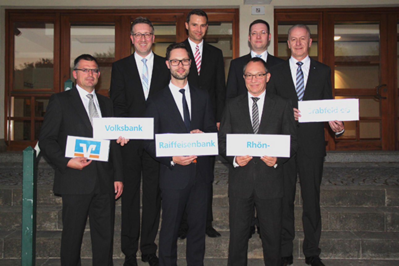 Auf einer Treppe stehen sieben Männer die Schilder mit der Aufschrift Volksbank Raiffeisenbank Rhön-Grabfeld eG