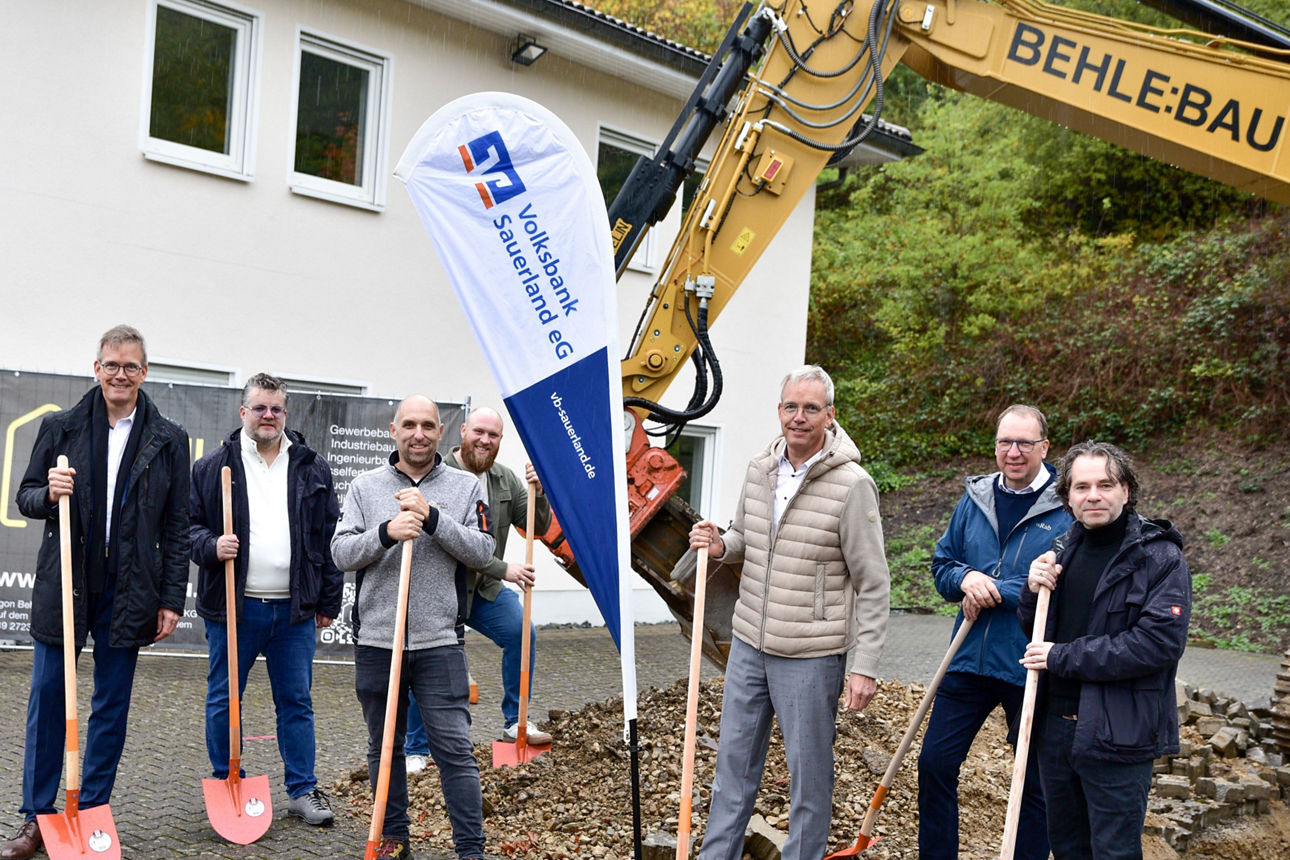 Eine Gruppe von Personen mit roten Schaufeln in den Händen, im Hintergrund ein gelber Bagger