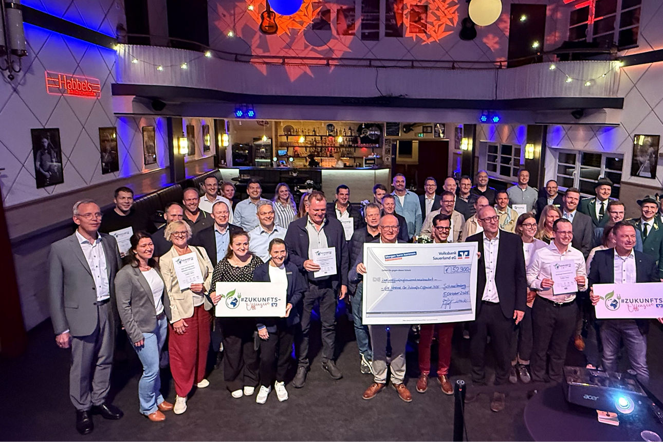 Eine große Gruppe von Menschen sind versammelt in einer Halle, einige halten Geld-Schecks in der Hand