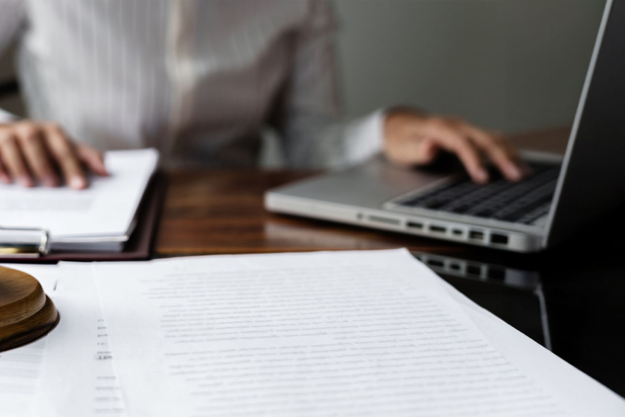 Holzhammer mit Unterlage liegt auf Formularen. Im Hintergrund ist eine Person zu sehen, welche am Laptop sitzt