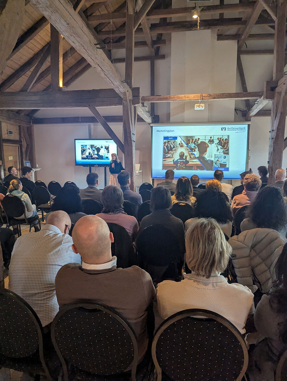 Ein großes Publikum hört einer Präsentation mit Fernseher und Leinwand zu.
