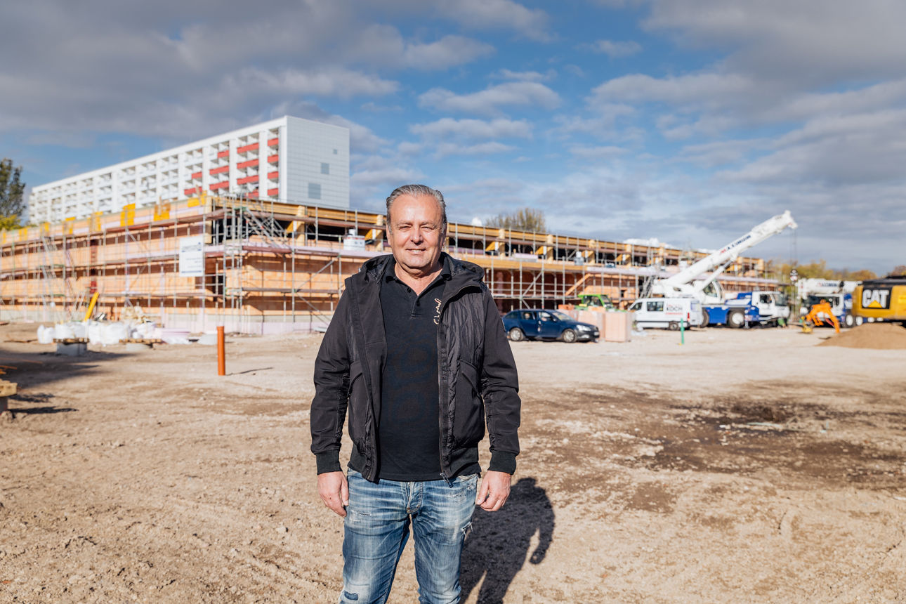 Karl‑Heinz Zehentner steht im Vordergrund einer Baustelle eines Fachmarktprojekts von „PZ Marktbau“. Im Hintergrund ist ein weit fortgeschrittener Rohbau eines Fachmarktzentrums zu sehen, der den aktuellen Stand der Bauarbeiten widerspiegelt