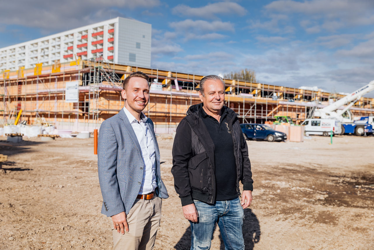 Berater Julian Trabert und Karl-Heinz Zehentner auf einer Baustelle vor einem im Bau befindlichen Fachmarktzentrum mit Gerüsten und Arbeitsfahrzeugen.