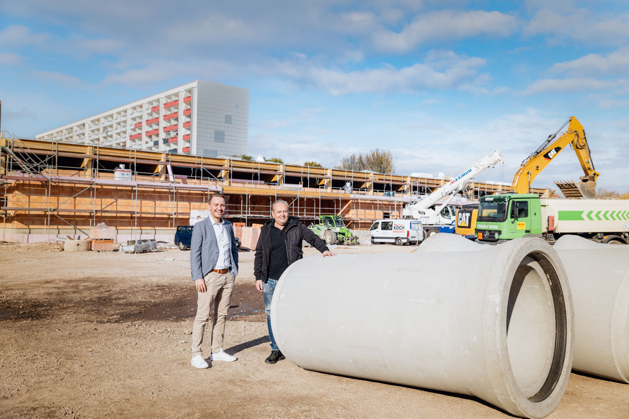 erater Julian Trabert und Karl‑Heinz Zehentner stehen auf der Baustelle eines Projekts von „PZ Marktbau“ vor einem angeordneten Betonfertigteil. Im Hintergrund sind Baufahrzeuge sowie der Rohbau des Gebäudes während der laufenden Bauphase zu sehen