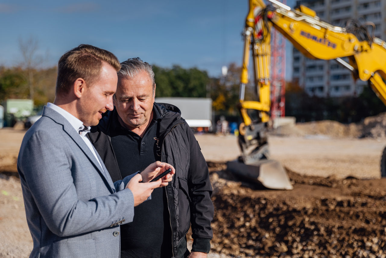 Berater Julian Trabert und Karl‑Heinz Zehentner stehen bei einer Abstimmung auf der Baustelle eines Immobilienprojekts von „PZ Marktbau“. Im Hintergrund sind Baugeräte sowie laufende Erdarbeiten im Bereich der Baustellenvorbereitung zu sehen