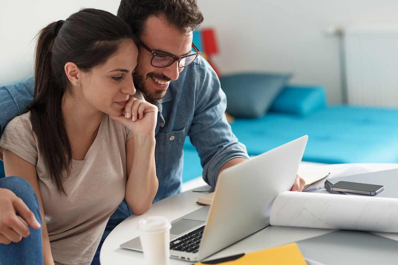 Paar am Laptop bei Online-Recherche zur Immobilienbewertung, Baupläne und Akten auf dem Tisch