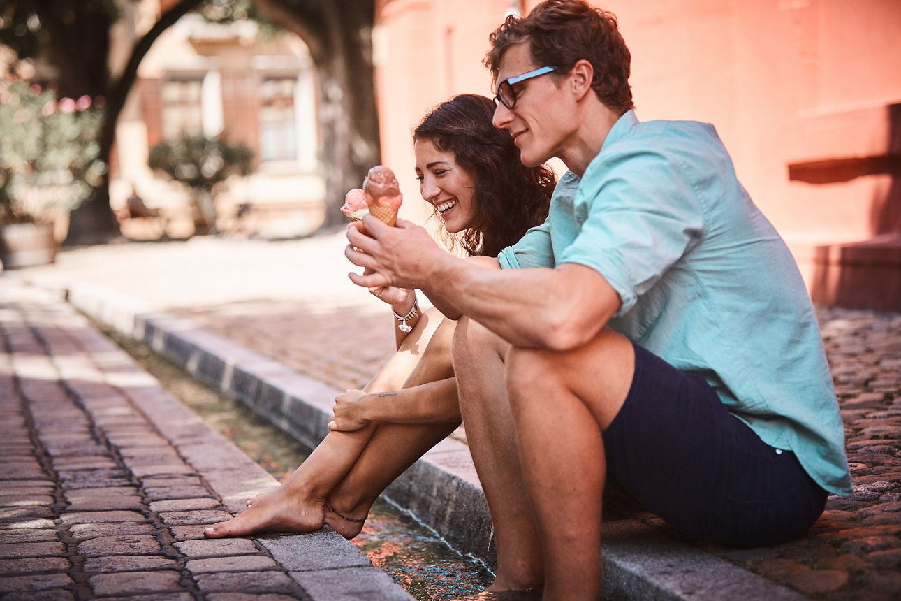 Eine Frau und ein Mann sitzen barfuß am Straßenrand und essen Eis in der Sonne.