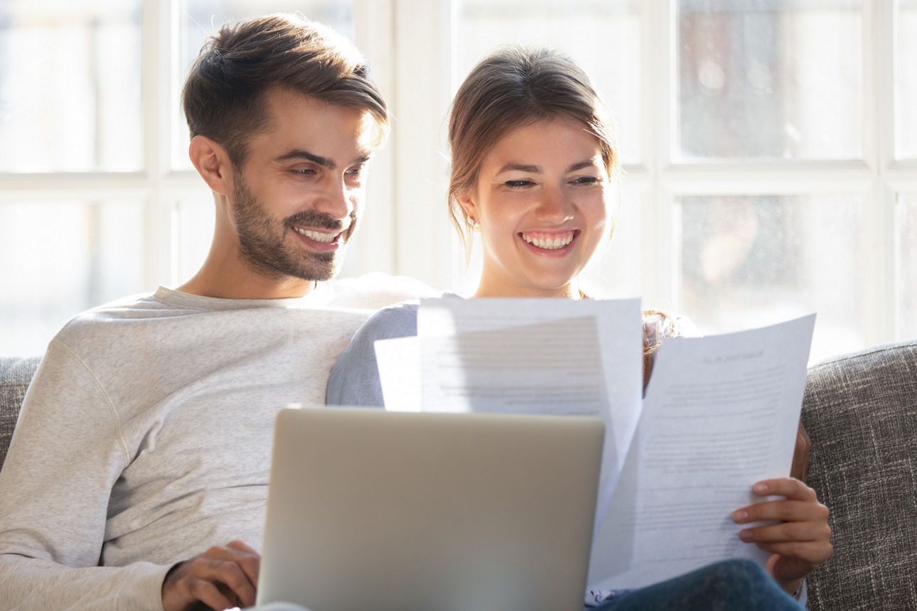 Mann und Frau sitzen nebeneinander auf dem Sofa und schauen auf Unterlagen. Er hält einen Laptop auf dem Schoß.