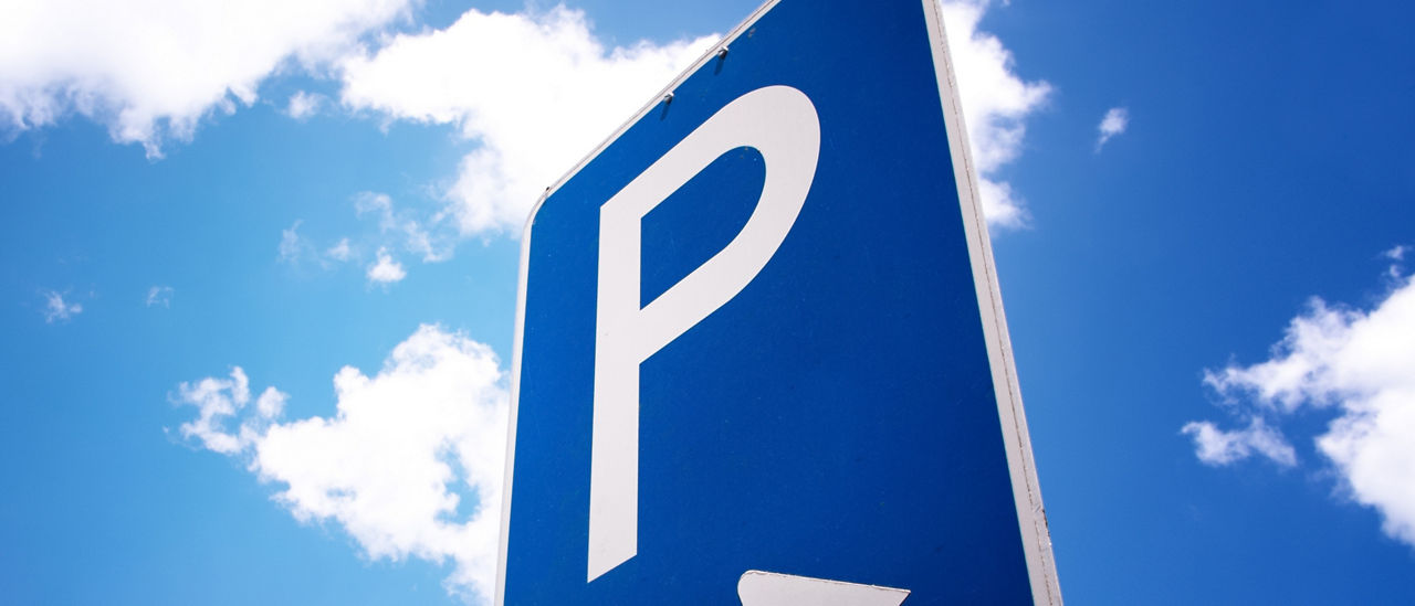 Ein Parkplatzschild mit Pfeil nach rechts steht vor strahlend blauem Himmel mit weißen Wolken. 