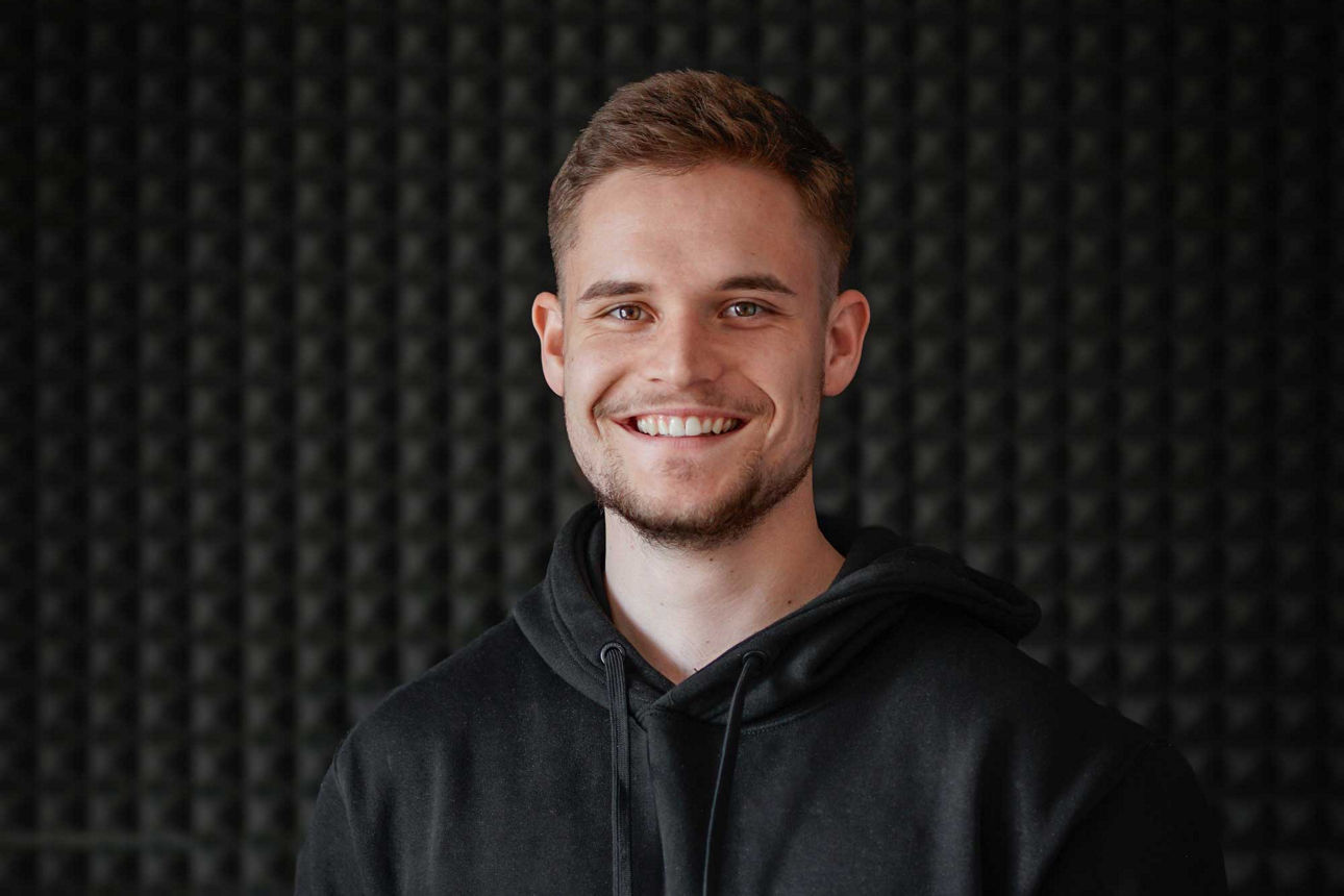 Mann in schwarzem Hoodie hält einen Laptop mit Apple-Logo in den Händen. Der Hintergrund besteht aus einer strukturierten, dunklen Wand.