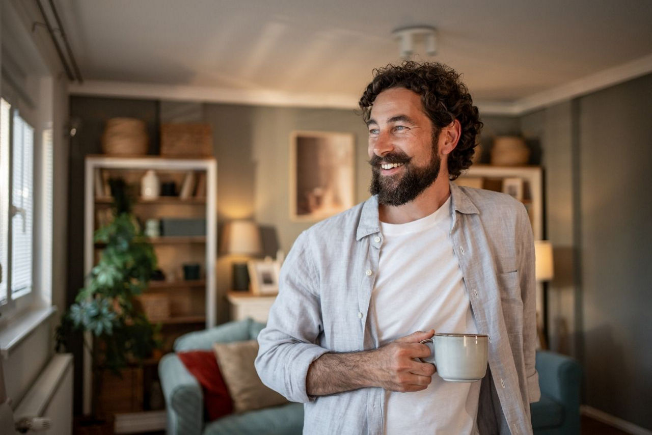 Mann mit Kaffeetasse in der Hand steht in seinem Wohnzimmer und sieht zufrieden aus