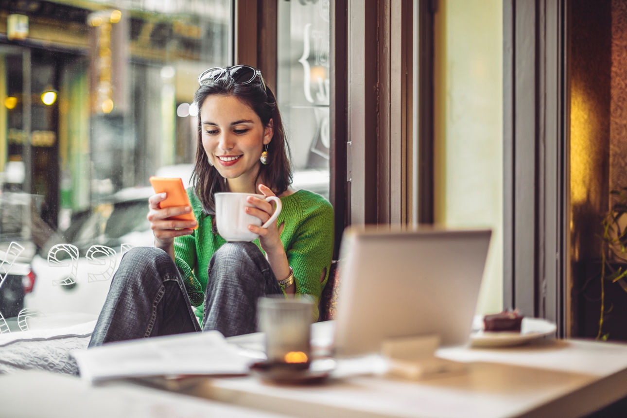 Person sitzt am Fenster in einem Café, schaut auf ein Smartphone und hält eine Kaffeetasse, Laptop liegt auf dem Tisch.