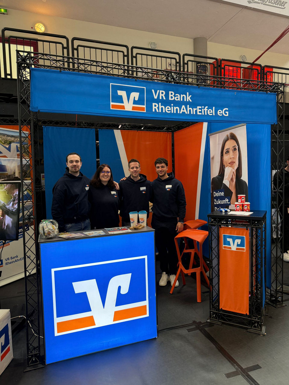 Messestand der VR Bank RheinAhrEifel eG mit vier Mitarbeitenden am Counter; Plakat: „Deine Zukunft. Die Bank.“