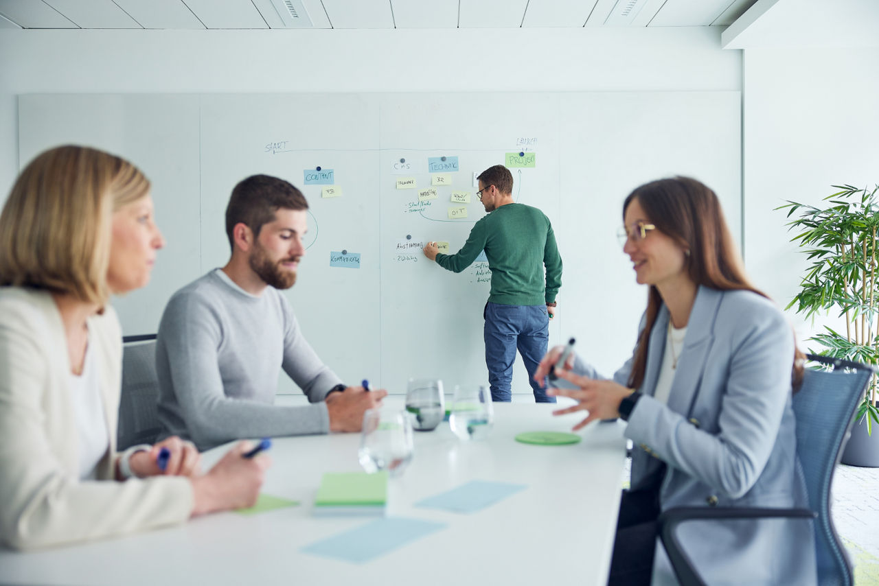 Drei Personen sitzen an Tisch und unterhalten sich im Hintergrund schreibt Mann etwas an ein Whiteboard