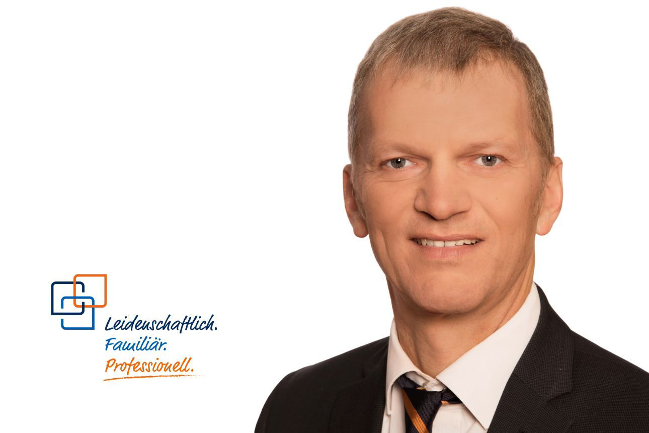 Peter Liebhart, ein Mann mit kurzen, grau-braunen Haaren, trägt einen dunklen Anzug mit einer gestreiften blau-orangenen Krawatte und blickt direkt in die Kamera.