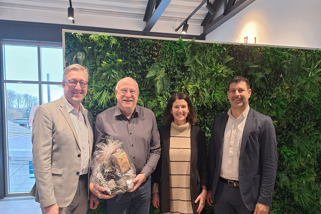 Das Bild zeigt vier Personen, drei Männer und eine Frau, die vor einer grünen Pflanzenwand stehen.  Der Mann in der Mitte häl