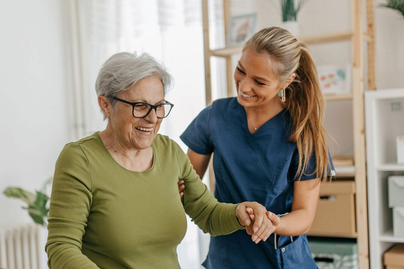 Pflegekraft unterstützt ältere Frau beim Gehen mit Gehstock im Wohnraum.