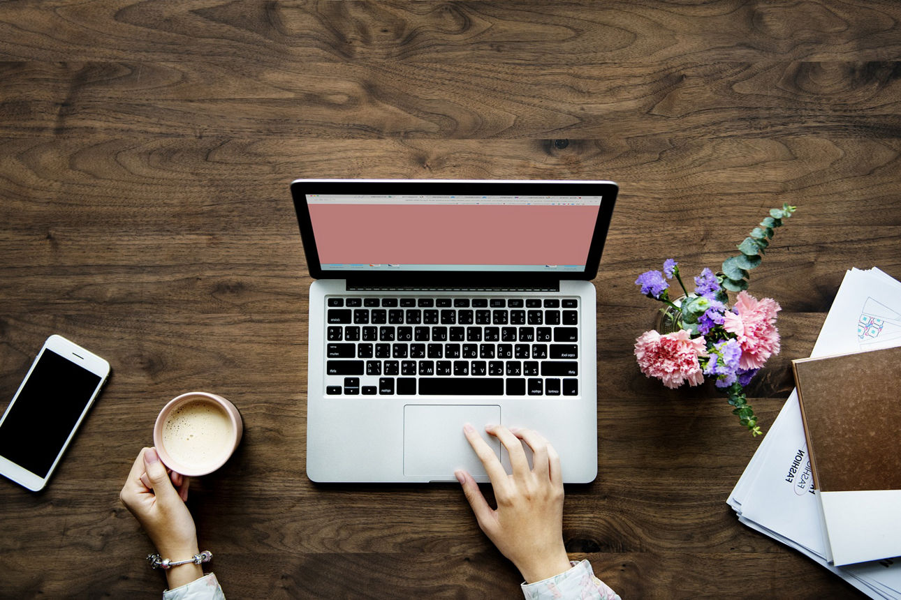 Blick auf einen Schreibtisch von oben, an dem eine Frau mit einer Tasse Kaffee vor dem Laptop sitzt 