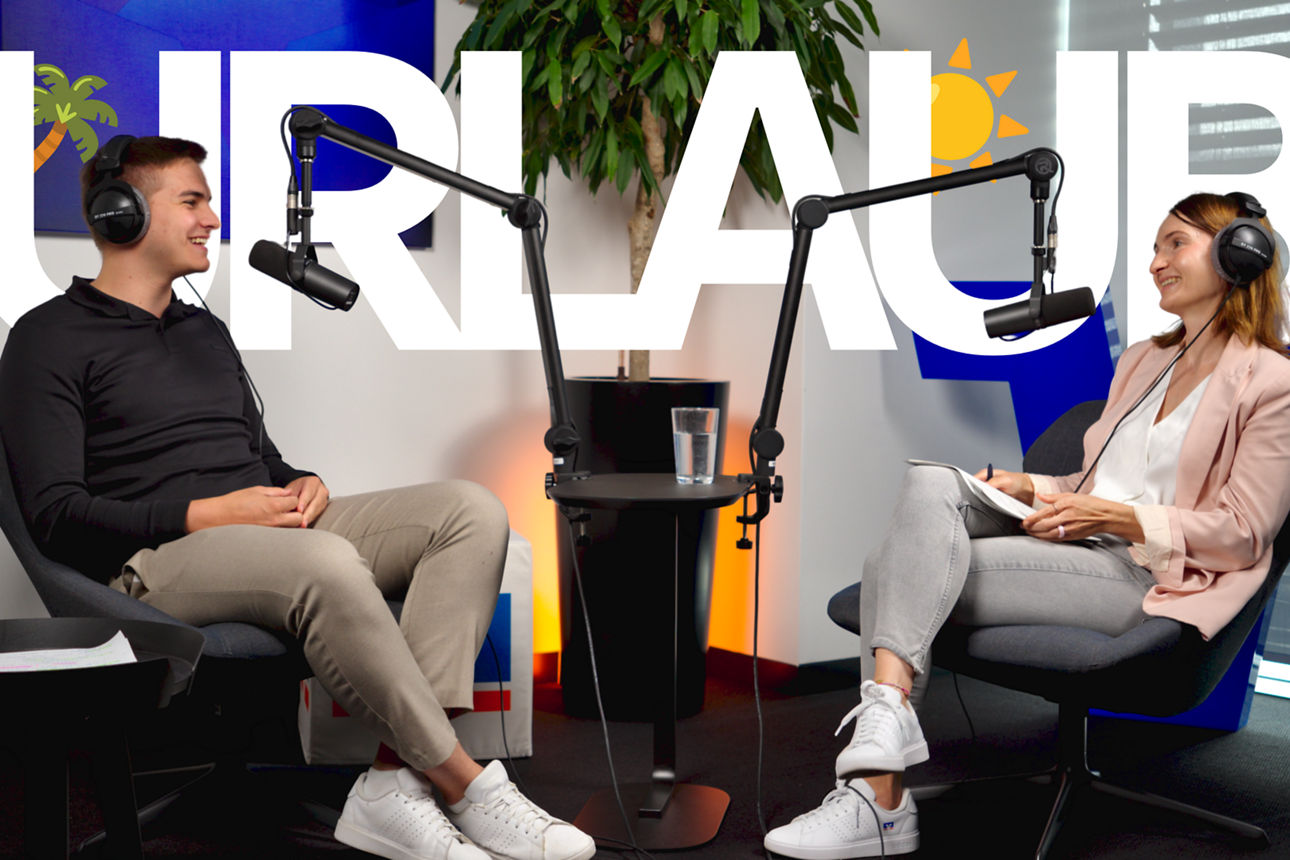 Zwei Personen sitzen sich in einem Podcast-Studio mit Mikrofonen gegenüber, im Hintergrund steht groß „URLAUB“.