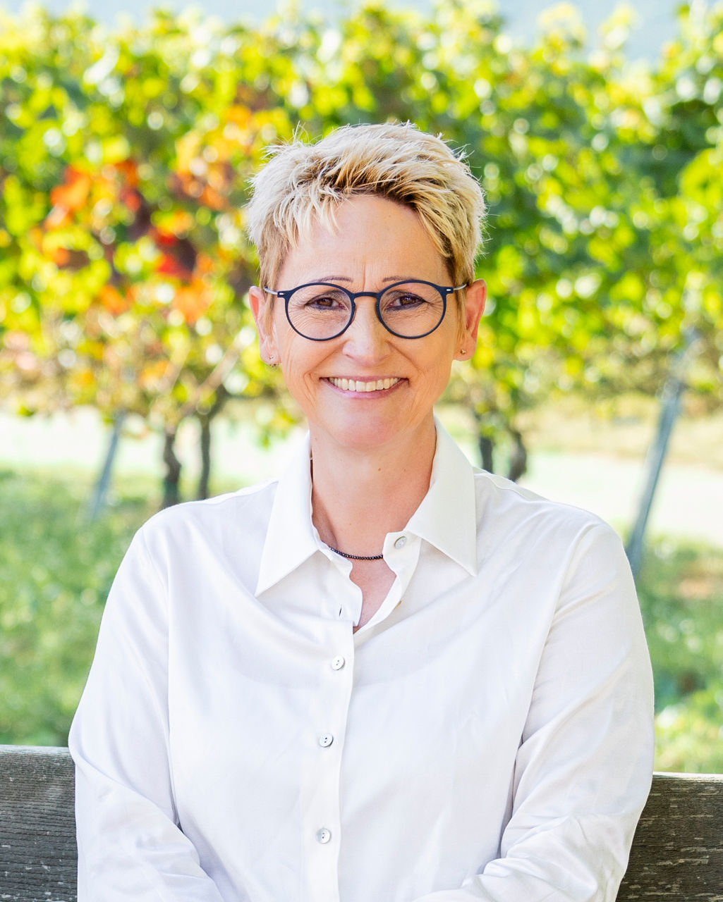 Lächelnde Frau mit Brille und weißer Bluse sitzt vor Weinreben im Freien.
