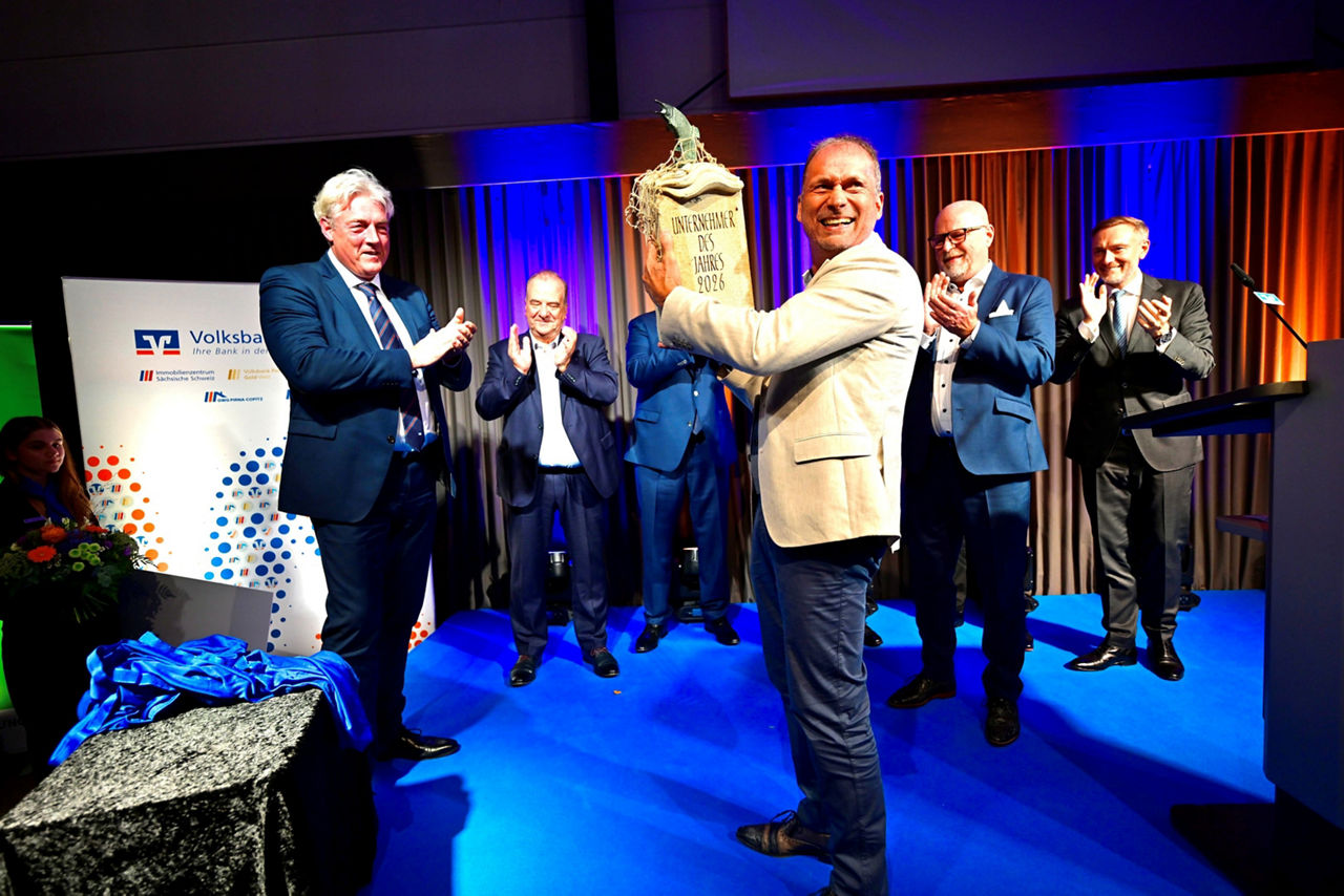 Mann im Anzug übergibt anderem Mann Pokal, beide lachen und freuen sich