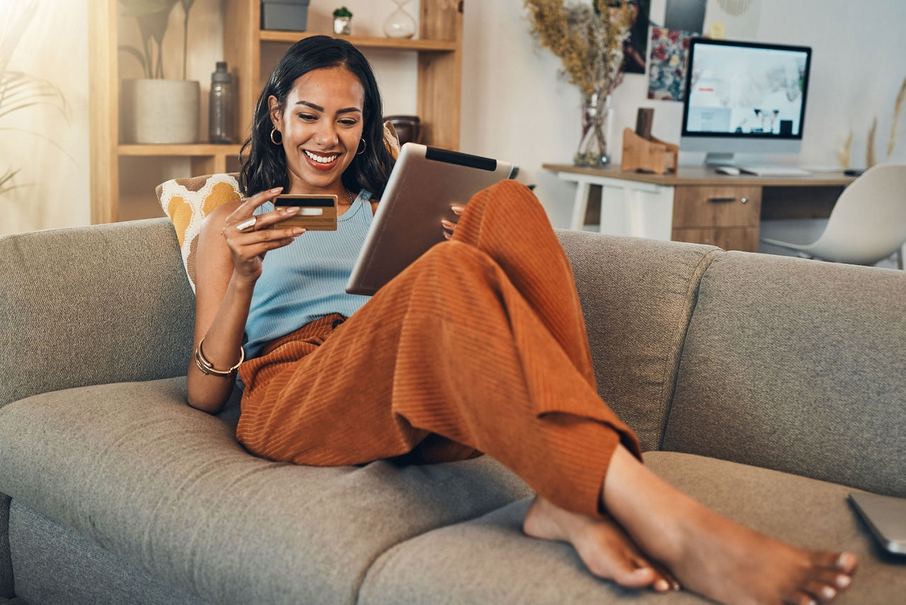 Junge Frau entspannt auf dem Sofa mit Tablet und Kreditkarte in der Hand.