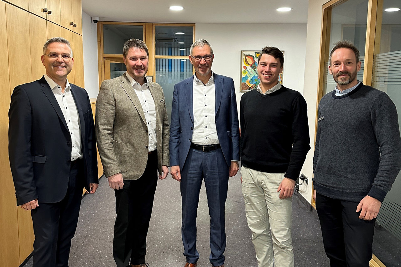 Fünf Männer stehen im Halbkreis in einem Bürogang und lächeln in die Kamera.