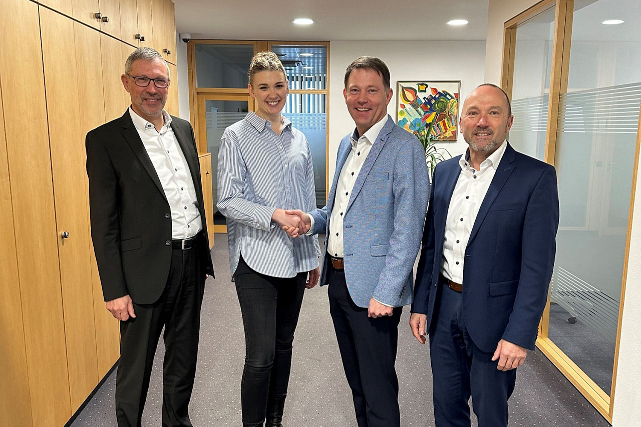 Drei Männer und eine Frau stehen in einem Bürogang. Die Frau und ein Mann stehen in der Mitte und schütteln sich die Hände.