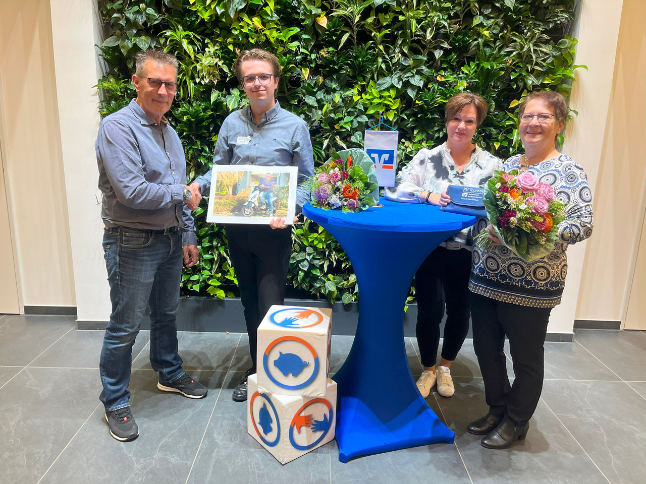 Vier Personen neben einem Stehtisch vor einer Pflanzenwand mit zwei Blumensträußen.