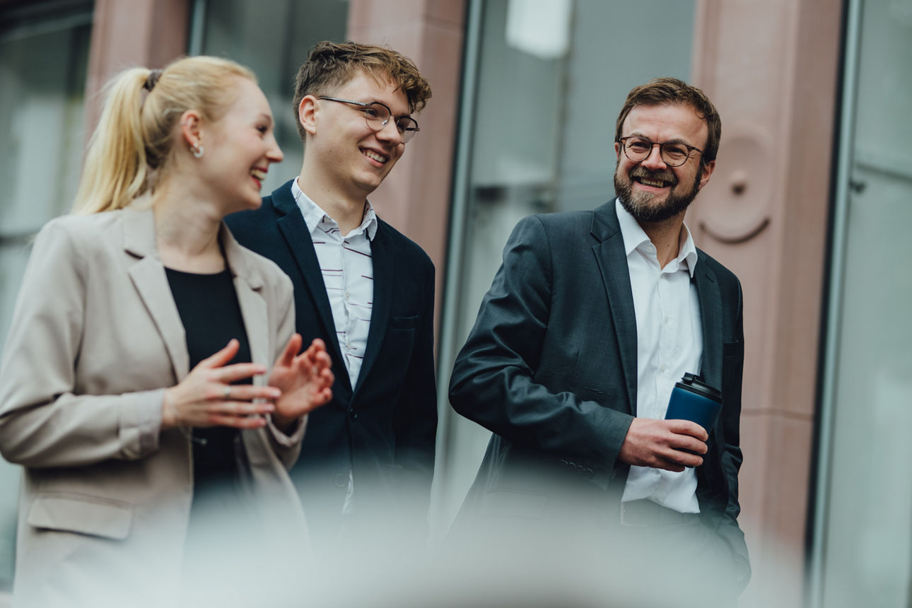 Drei Mitarbeitende laufen vor einem Gebäude lang und unterhalten sich