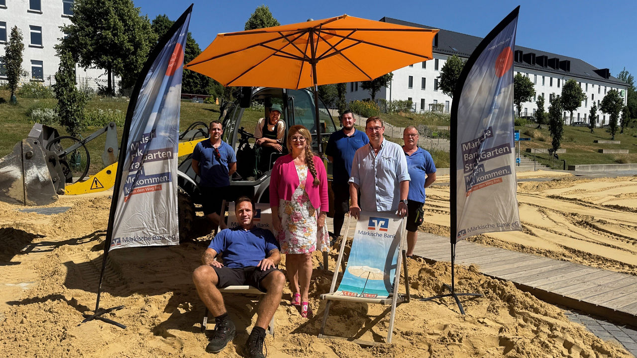 Mehrere Personen stehen und sitzen unter einem orangefarbenen Sonnenschirm auf einem Sandplatz.