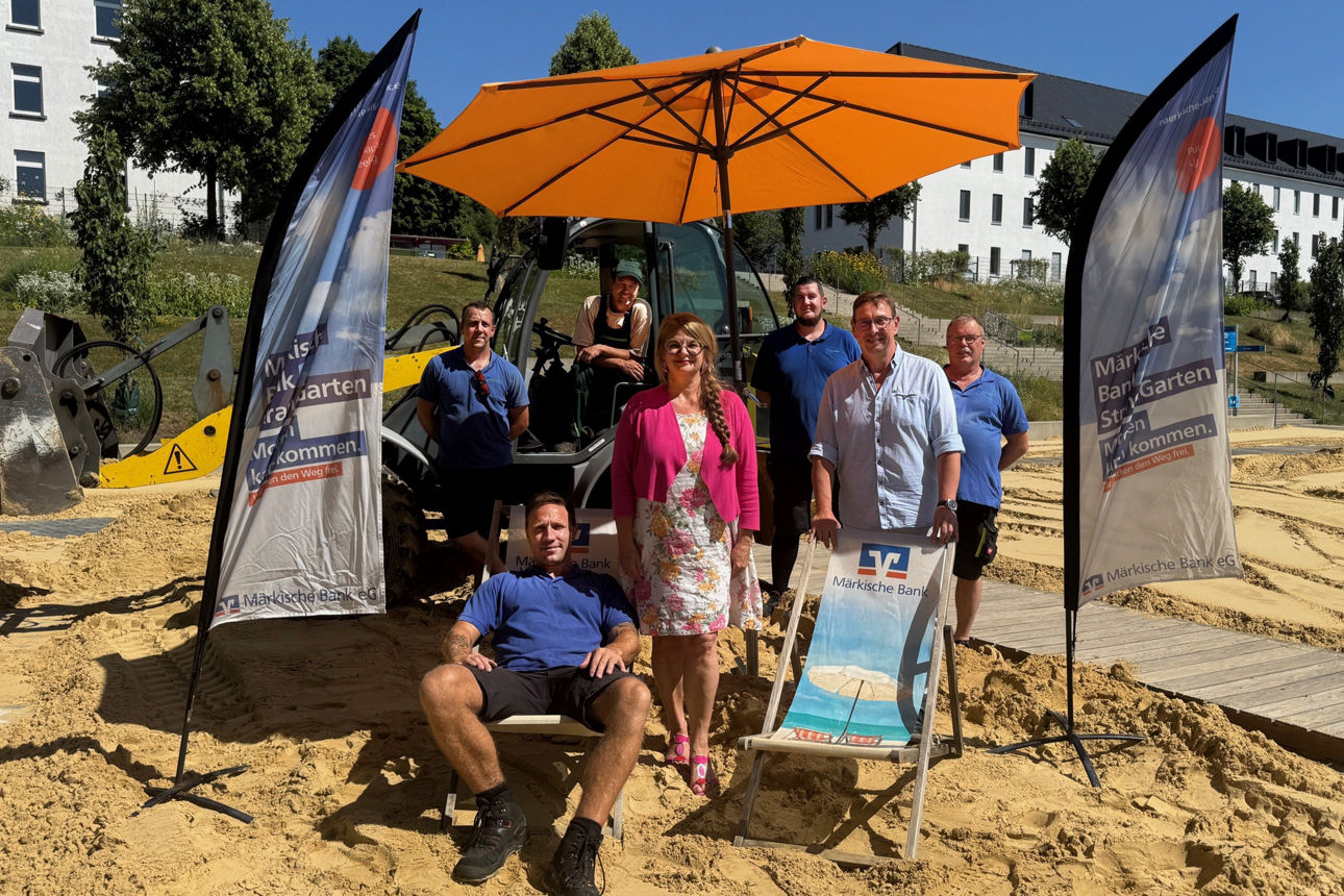Mehrere Personen stehen und sitzen unter einem orangefarbenen Sonnenschirm auf einem Sandplatz.