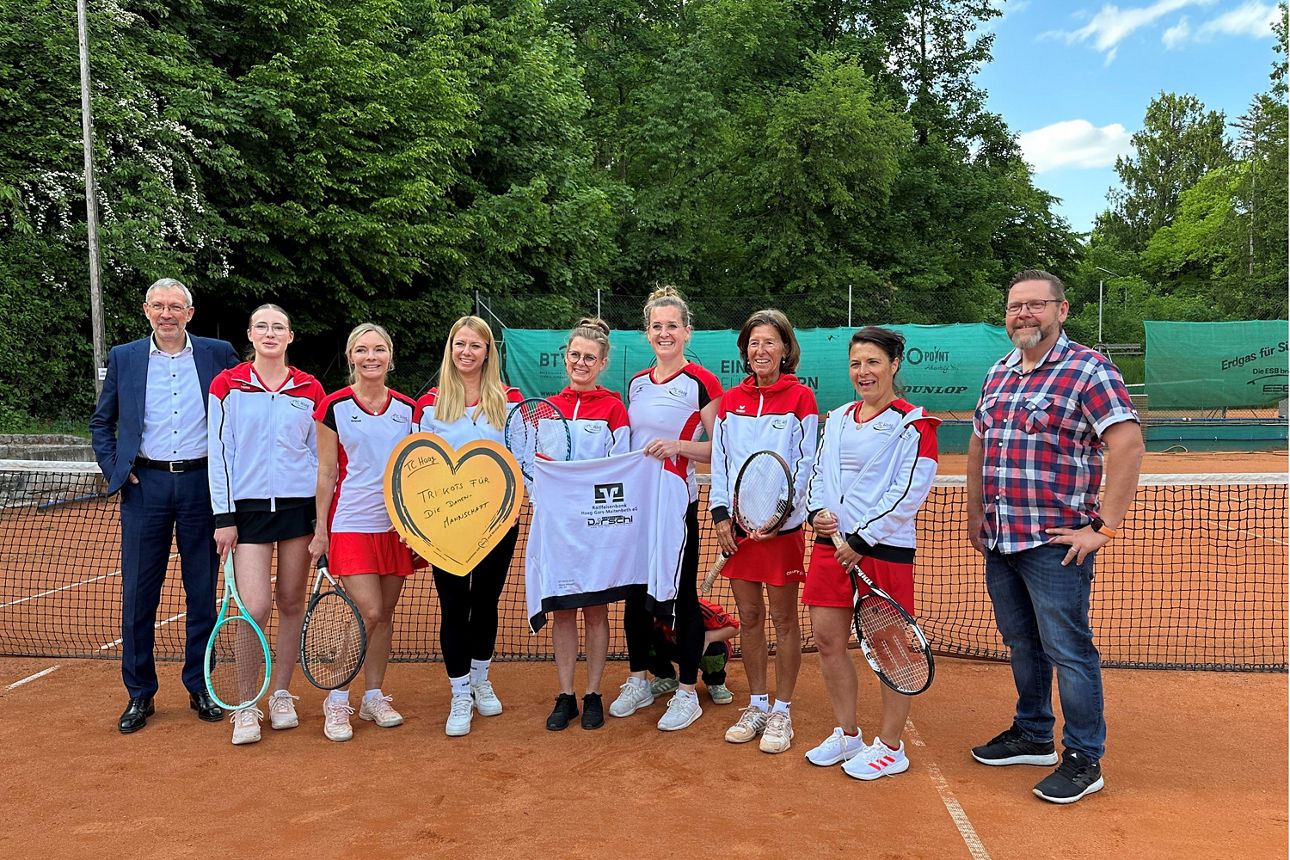 Neun Personen, darunter mehrere in Tenniskleidung, posieren auf einem Sandplatz mit einer Urkunde