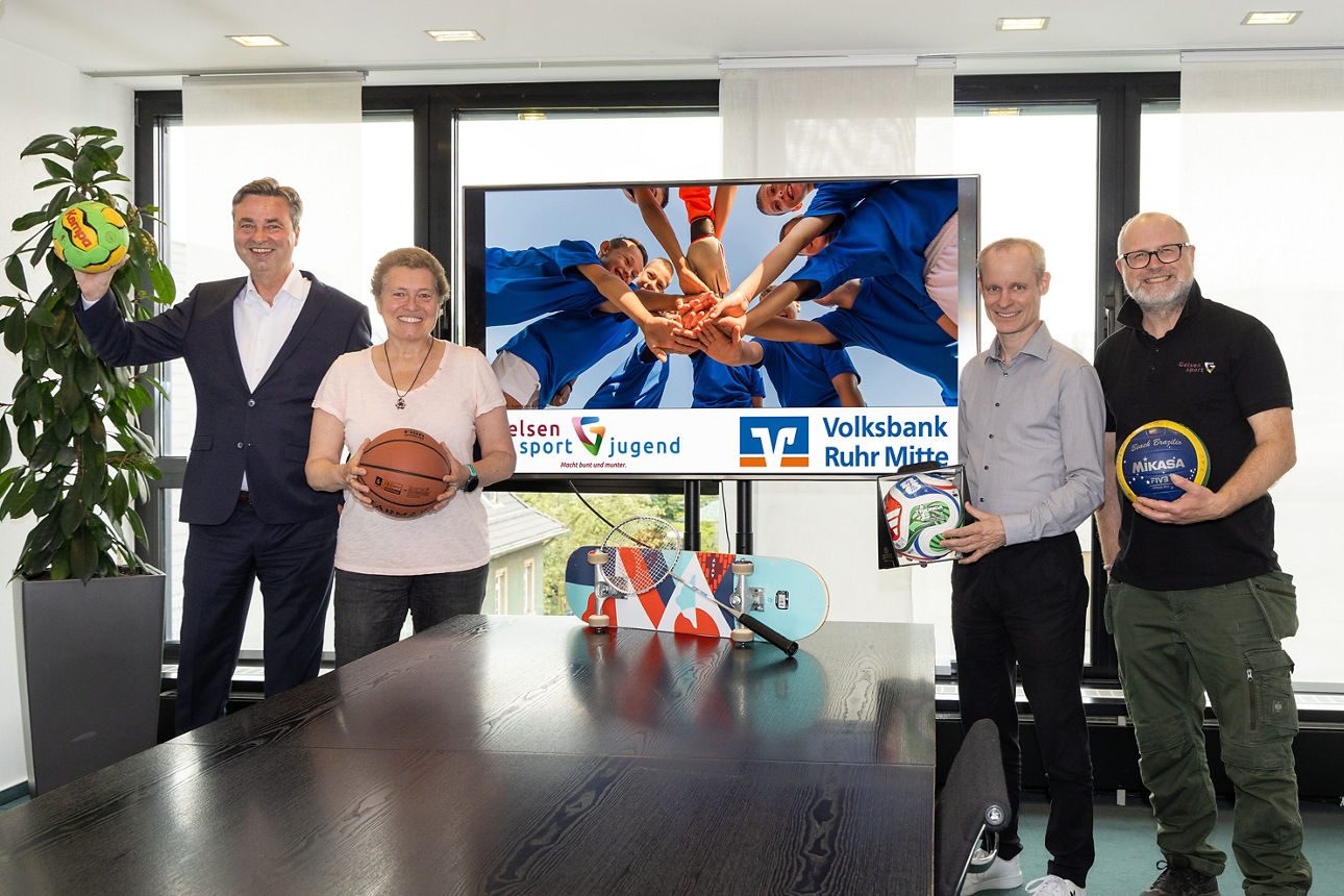 Vier Personen stehen in einem hellen Raum neben einem großen Bildschirm und halten verschiedene Sportbälle in den Händen. Auf dem Bildschirm ist ein Bild von Kindern mit ausgestreckten Händen sowie die Logos von Gelsensport Jugend und der Volksbank Ruhr Mitte zu sehen. Im Vordergrund stehen auf einem Tisch weitere Sportgeräte wie ein Volleyball, ein Fußball, ein Badmintonschläger und ein Skateboard. Die Szene wirkt wie eine gemeinsame Präsentation oder Kampagne rund um Sport und Engagement.